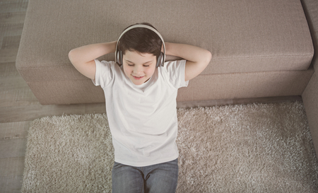 Relaxed boy is listening to music from headphones. He is lying on floor while leaning head on sofa. Top viewの写真素材