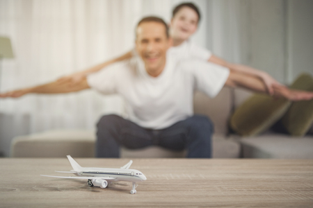 Dreaming about family travel. Focus on small toy plane on table. Happy father and son are stretching their arms as wings and laughing at home. They are as if flying on backgroundの写真素材