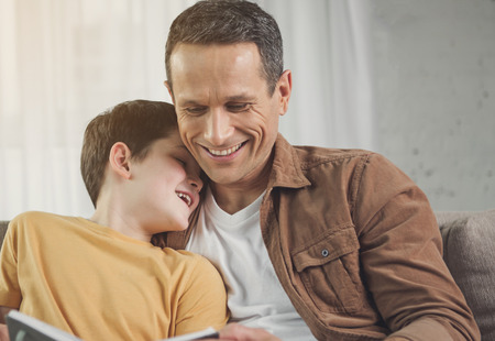 Portrait of happy father reading fairy tale for his lovely son. Boy is leaning head on male shoulder and laughingの写真素材