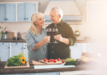 Portrait of positive senior married couple drinking wine while cooking romantic dinner together. They are cuddling and laughingの写真素材