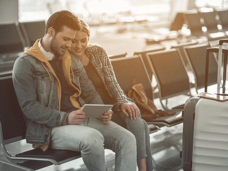 Outgoing male telling with satisfied woman while watching at electronic tablet. Glad couple using gadget before tripの写真素材