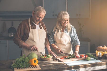 Joyful senior pensioners cooking together at home. Man is mixing salad while woman is cutting vegetables. They are wearing headphonesの写真素材