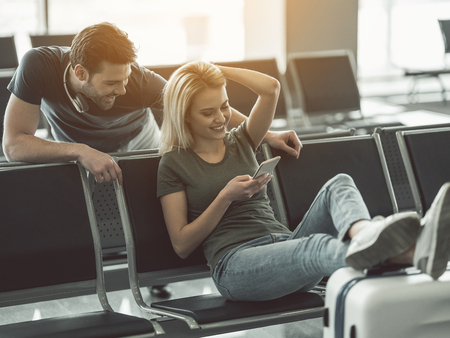Outgoing man and happy woman looking at phone while talking together in airport. Cheerful girl and smiling male using appliance conceptの写真素材