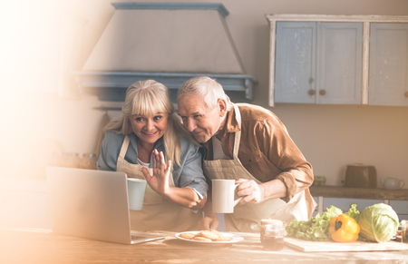 Hello. Portrait of excited old grandparents talking with family by internet. Woman is waving hand to screen and smiling. Man is standing in kitchen and holding cupの写真素材