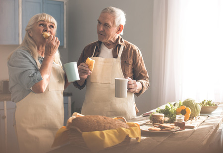 Joyful mature couple is tasting sweet self-baked buns in kitchen. They are standing and drinking teaの写真素材