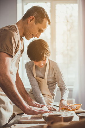 Father standing in apron and unrolling soft dough on table while his kid observing process. They are smilingの写真素材