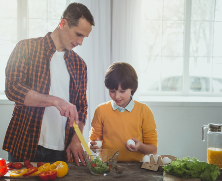 Parent and child standing at the kitchen table. Father pointing at bowl with greens, attentive kid holding eggsの写真素材