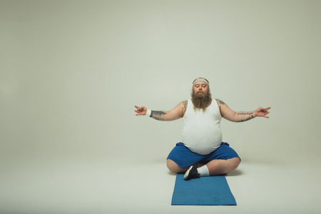Full length portrait of calm fat man meditating with concentration. He is sitting on mat in lotus position. Copy spaceの写真素材