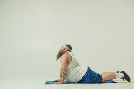 Calm fat man is doing healthy exercise with concentration. He is lying on mat while stretching body behind. Copy spaceの写真素材