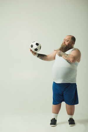 Stay still. Cheerful obese guy is pointing at a football with command while holding a toy in hand. Copy spaceの写真素材