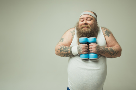 Waist up portrait of happy fat guy is hugging dumbbells and smiling. His eyes are closed with pleasure. Isolated and copy spaceの写真素材