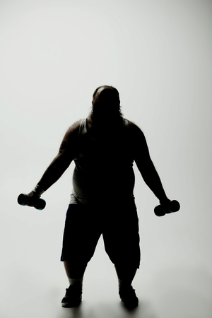 Dark silhouette of strained fat man exercising with weights while standingの写真素材