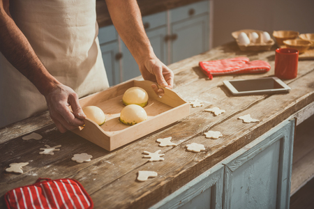 Man hands putting tray with tasty dessert on kitchen table. Uncooked dough and tablet are on desk. Close upの写真素材
