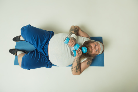 Top view of scared fat man is staring at camera with shock. He is lying on sporty mat and holding dumbbellsの写真素材