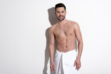 Portrait of cheerful young bearded man in towel is standing against white wall and looking at camera with joy. Copy space in the left side.の写真素材