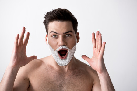 Portrait of surprised topless guy with shaving cream on face is standing and looking at camera with...