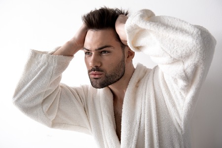 Portrait of confident young bearded man in white bathrobe is standing and touching his hair. He is...