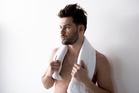 Young sleepy bearded man with tousled hair is standing against light wall and holding white towel...