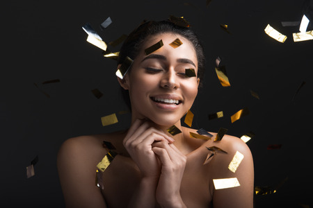 Portrait of good looking woman with eyes closed standing in cloud of confetti. Isolated on black...