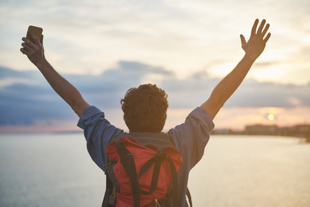 Back view of happy male tourist stretching arms up while watching at sunset near the ocean. Feeling freedom conceptの写真素材