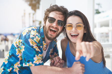 Waist up portrait of joyful young loving couple enjoying sea view at the beach. Woman is pointing finger forward and smiling with excitementの写真素材