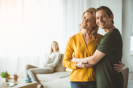 Waist up portrait of happy married couple are standing and embracing. Pregnant surrogate woman is sitting on sofa on backgroundの写真素材