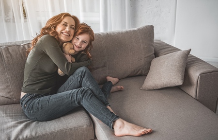 Love you so much. Full length portrait of delighted red-haired woman hugging her daughter tightly on couch. Little girl is holding her teddy bear with smile and joyの写真素材