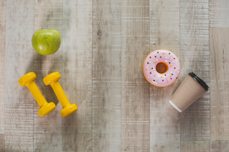 Sport and health versus harmful habits. Top view of green apple and yellow dumbbells lying opposite to coffee cup with donuts on wooden floor. Close up