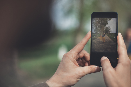 Close up of male hands holding smartphone. Man is photographing woman standing on the road in the nature. Focus on screen の写真素材