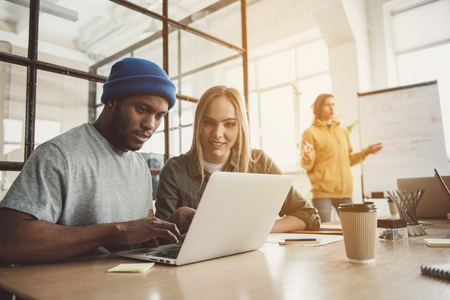 Portrait of happy lady and optimistic man typing in gadget while sitting at table. Colleague telling near board. Working process conceptの写真素材
