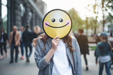 Be happy. Waist up portrait of trendy lady with round cheerful smile in her hand hiding her face. She is standing in the street and a lot of people on the backgroundの写真素材