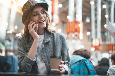 Smiling lady in trendy hat is chatting on smartphone in a cafe. She is drinking hot drink during conversationの写真素材