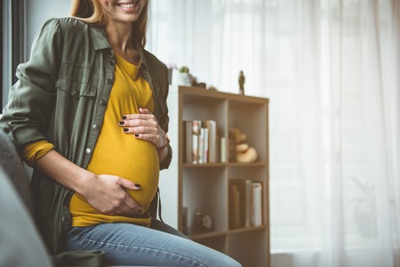 Low angle of joyful expectant mother is sitting on windowsill and embracing her belly. She is smiling with happiness. Copy space in right sideの写真素材