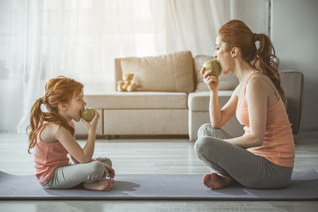 Profile of woman and kid after aerobics. They are sitting in front of each other on the floor and biting green apples with smileの写真素材