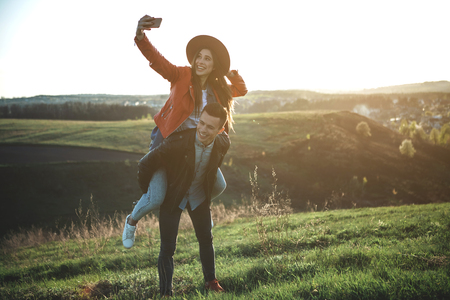 Forever young. Full length of laughing female and male surrounded by wonderful green hills. Woman is making selfie on back of boyfriend. Copy space in right sideの写真素材