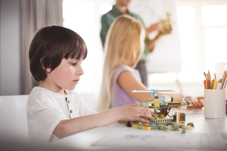 Side view orderly kid making helicopter at table. He sitting opposite girl. She talking with...