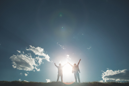 Summer of love. Full length of girl and boy rejoicing amazing sun on green meadow. They are standing close to each other with hands up facing beautiful blue skyの写真素材