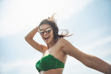 Happy day. Low angle portrait of young cheerful attractive woman in sunglasses and swimsuit is standing outdoors and enjoying summer vacations abroad. She is looking at camera with joy. Travel conceptの写真素材