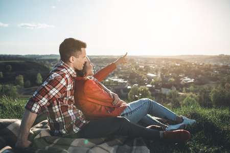 Look there. Full length side view of man and woman sitting and looking at beautiful summer sky. Delighted girl is pointing at shining sun. They are bonding to each other and smilingの写真素材