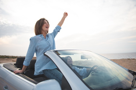 Full of happiness. Two overjoyed young attractive women are driving car while travelling along seashore. Girl is standing in cabriolet with open roof. She is feeling contentment with raised handの写真素材