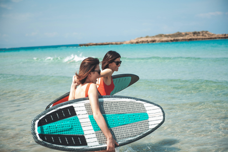 Side view of two happy young female friends in sunglasses are carrying their surfboards while wading into sea to surf. They are enjoying summer holiday. They are ready to have fun. Vacation conceptの写真素材
