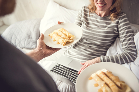 Top view focus on white plate with sweet rolls with topping. Gentle man is giving breakfast to soulmate while she is sitting in bed with laptop. Grateful woman is taking dish and smiling with delightの写真素材