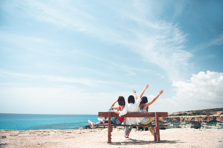 Fun together. Full length back view of three cheerful girls are sitting on bench and looking at azure ocean. They are expressing gladness with raised hands. Summer trip concept and copy spaceの写真素材