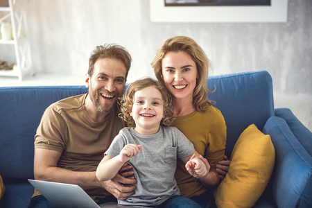 Waist up portrait of cheerful parents sitting with son near laptop. They are laughing and staring at camera from comfortable couch. Boy is staying on mother lap while couple is embracingの写真素材