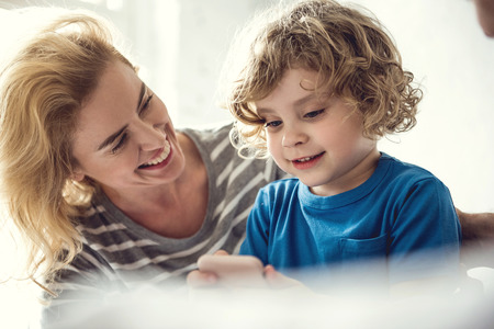Cheerful mom is enjoying time with little boy while he is using mobile phone. Cute kid is...