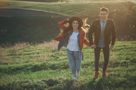 Full length of happy young people holding hands and flipping on meadow in summer evening. Girl is holding hat. Copy space in left sideの写真素材