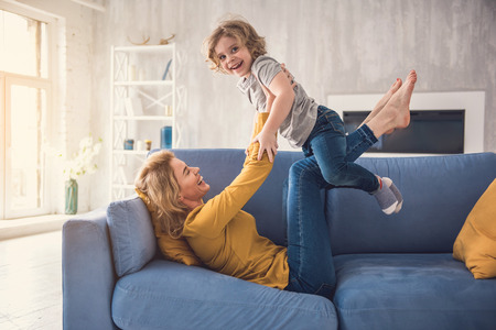 Mom and child are laughing on divan while spending time together. Woman is swinging and raising kid on her legs while lying on couch. Boy is holding parent arms and looking at camera with excitementの写真素材