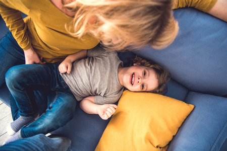 Top view of laughing son spending great time and looking at camera with joy. He is lying on sofa near his mother who is tenderly watching him. Parent is entertaining kid by kindly fondlingの写真素材