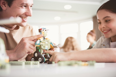 Cheerful man holding toy in hand. Beaming kid pointing at it. They situating at deskの写真素材