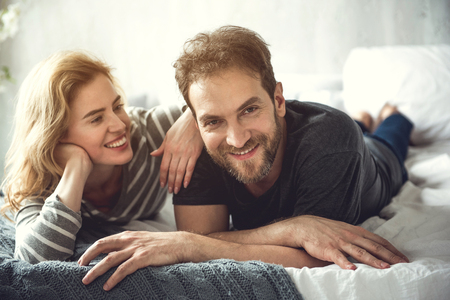 Cheerful bearded man is lying with soulmate on bed. He is looking at camera while woman is tenderly bonding him. Happy relationships conceptの写真素材